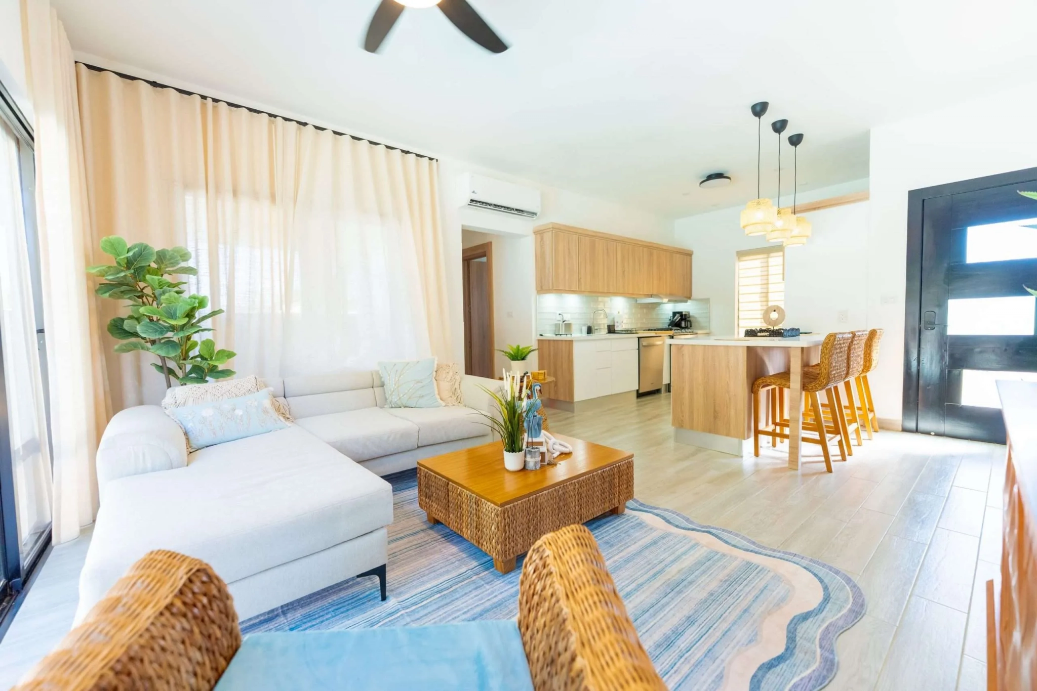 Bright living space with a white sofa, wicker chairs, and green plants, opening to a modern kitchen island in the background.