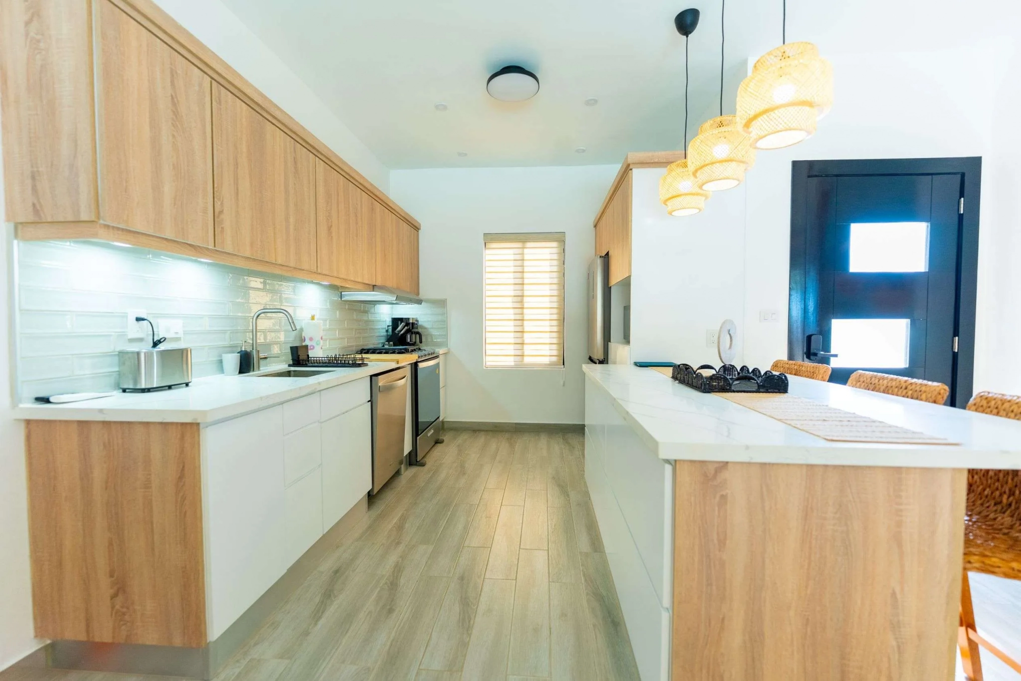 Bright modern kitchen with light wood cabinets, white countertops, stainless steel appliances, and woven pendant lights above an island.