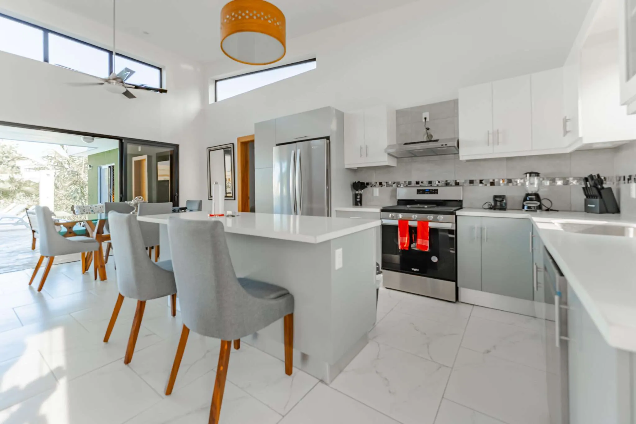 Bright modern kitchen with white cabinets, stainless steel appliances, and a large island with gray stools and wooden legs.