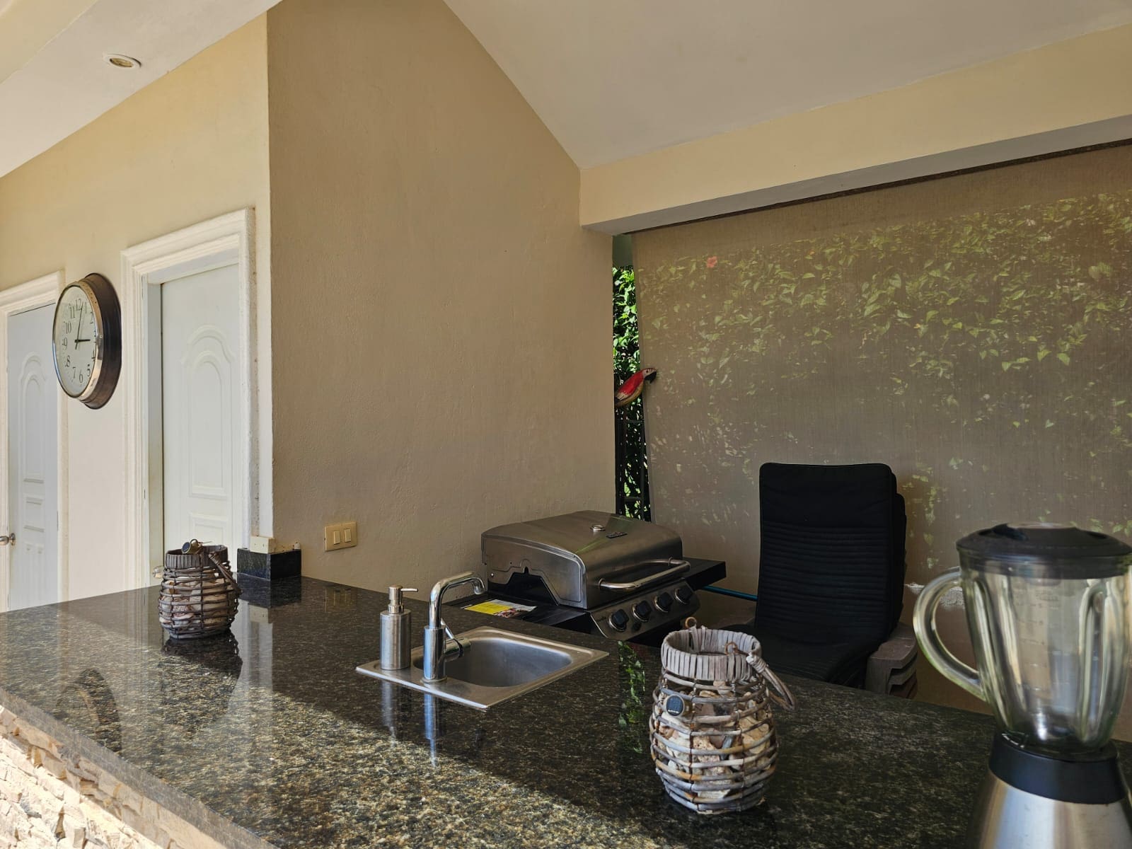 Outdoor kitchen bar with granite counter, built-in grill, sink, blender, and wicker lanterns; beige walls and a black chair nearby, with a red bird peeking through a gap behind.
