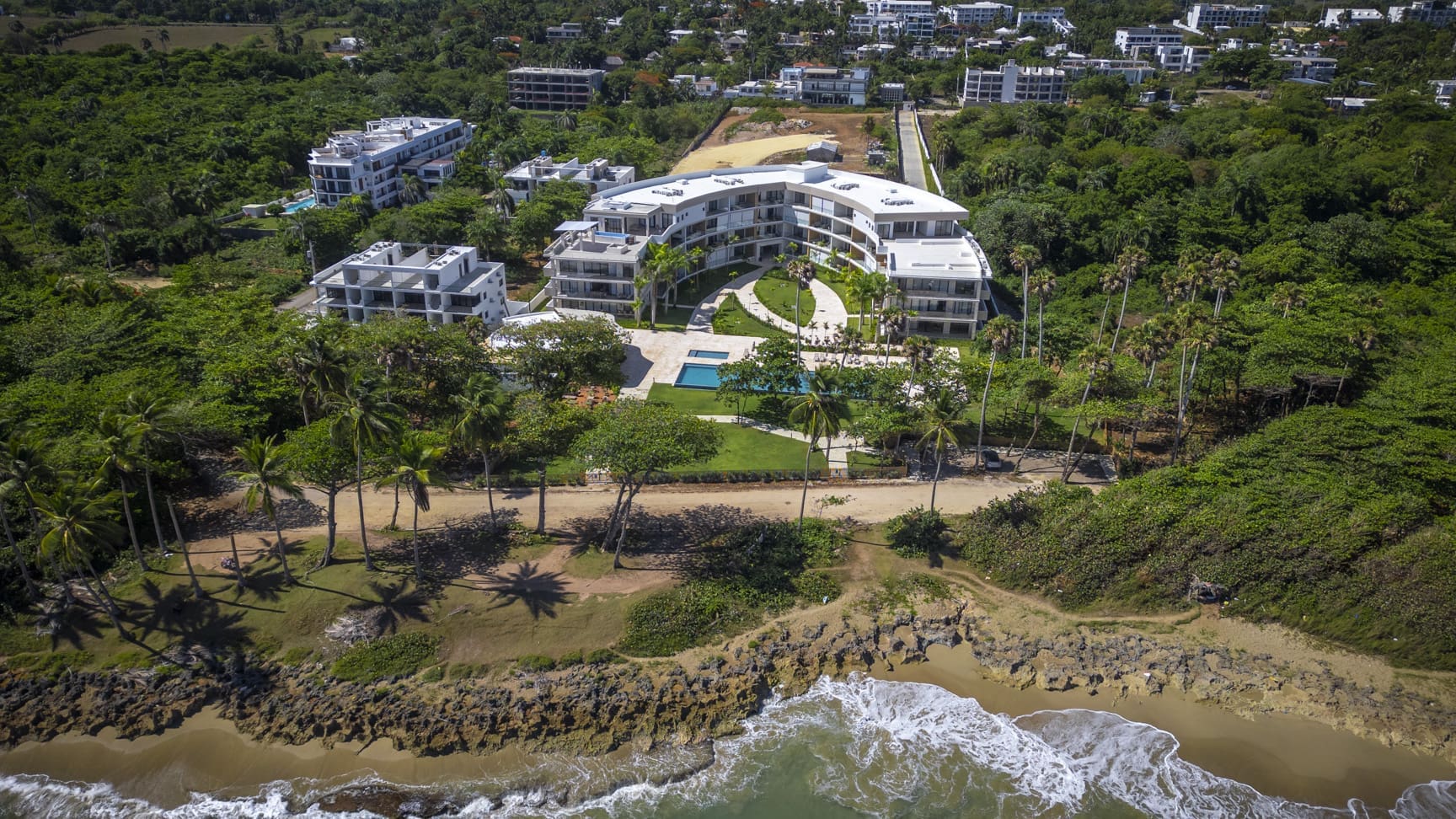 Aerial view of a modern curved resort complex with pools and palm trees on a tropical coastline.