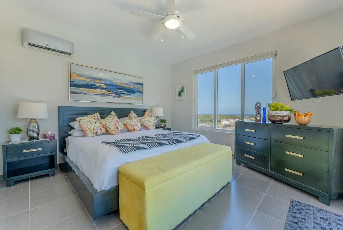 buyDRproperty Modern bedroom with king-size bed, yellow bench, and large window view Sosua Real Estate